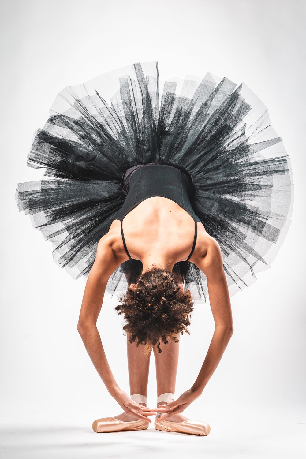 Ballet dancer in black tutu bends forward in graceful pose while wearing pink pointe shoes on white background.