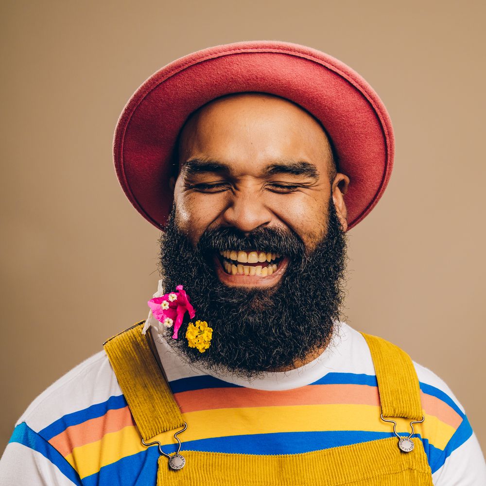 Cheerful person in red hat and striped shirt wearing overalls and sporting a full dark beard laughs joyfully.