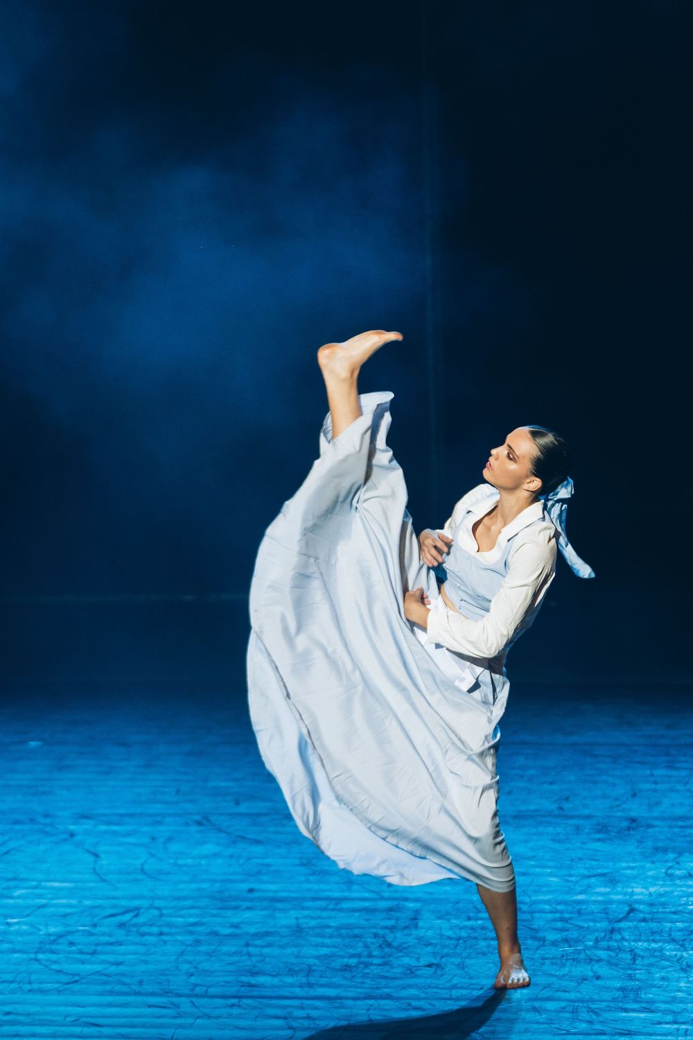 Graceful dancer in flowing white dress performs dramatic high kick against dark blue background on stage.