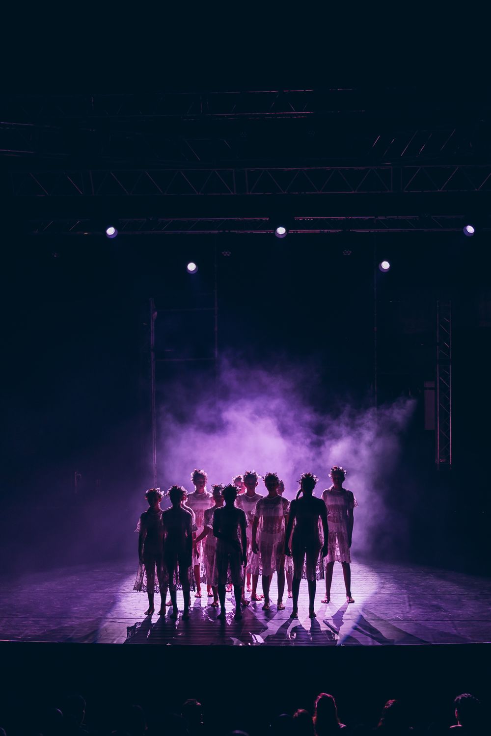 Silhouetted performers on a dark stage with dramatic purple smoke effects and overhead stage lighting.