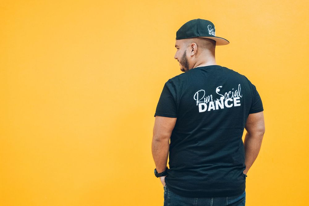 Back view of someone wearing a black t-shirt with dance text standing against a bright yellow wall.