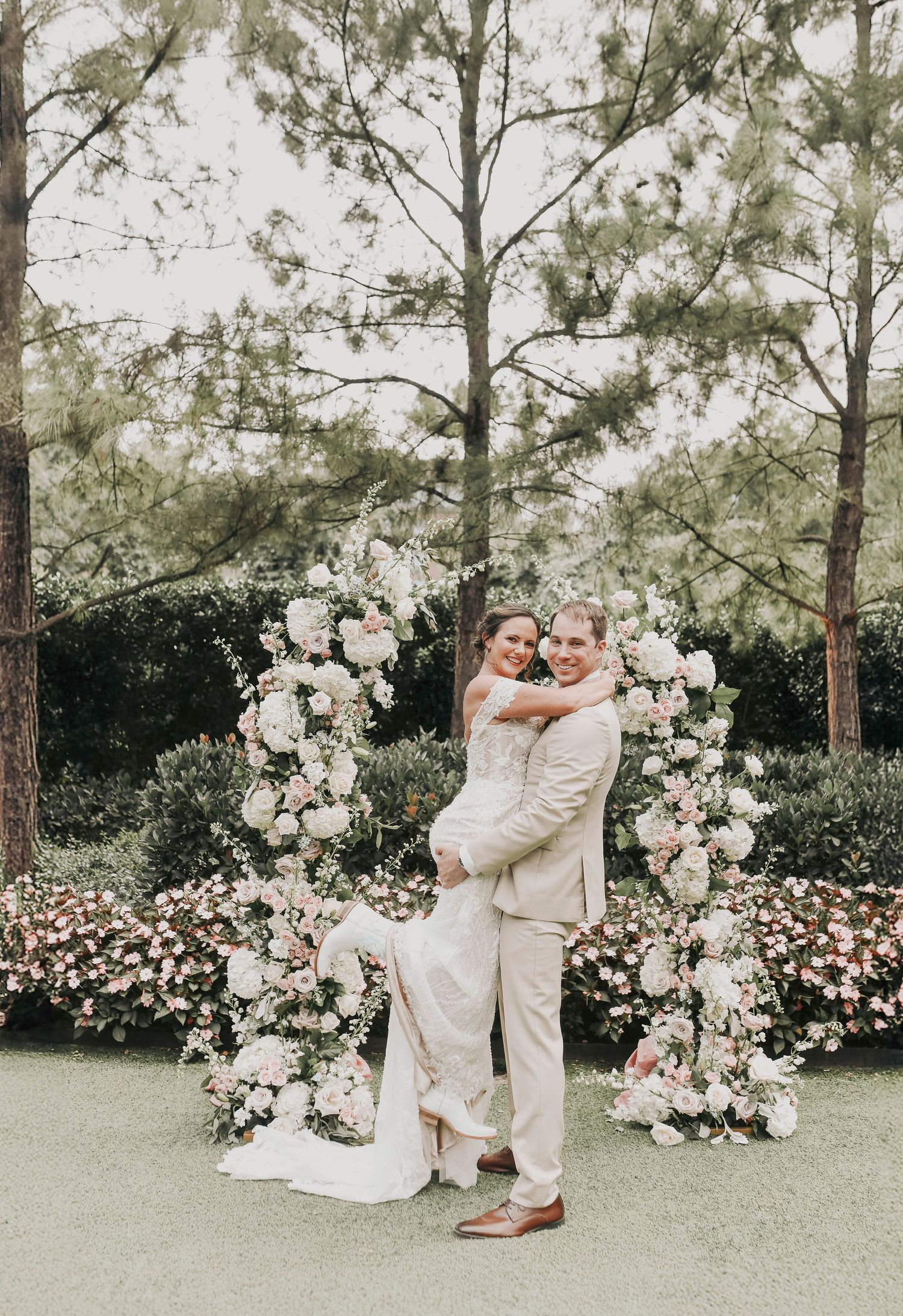 happy bride is lifted up by her groom as she shows off her white cowgirls boots after their wedding ceremony in the garden of the drover