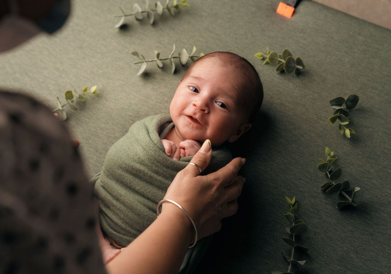 A small infant wrapped in a sage green swaddle lies on a carpet surrounded by eucalyptus leaves during a photo session.