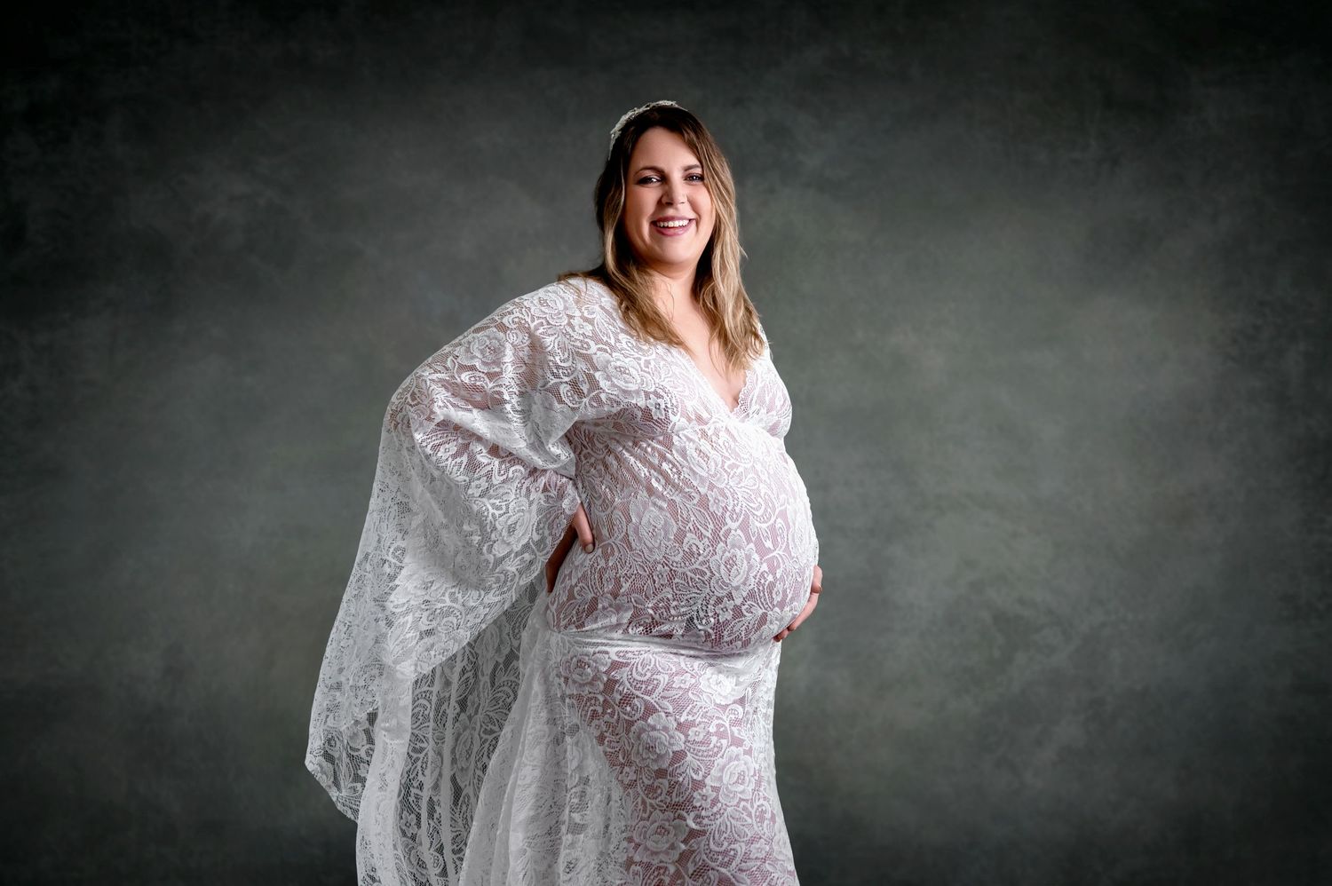 Expecting person poses in white lace maternity gown against dark studio backdrop with atmospheric lighting.