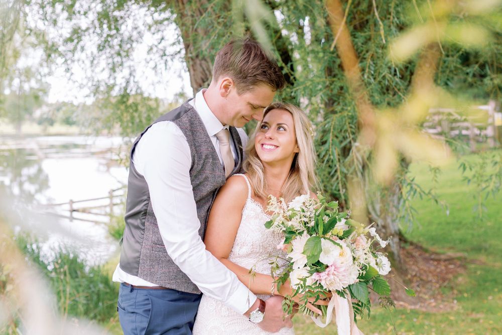 Joyful couple in wedding attire embrace near a lake with a romantic floral bouquet in a garden setting.