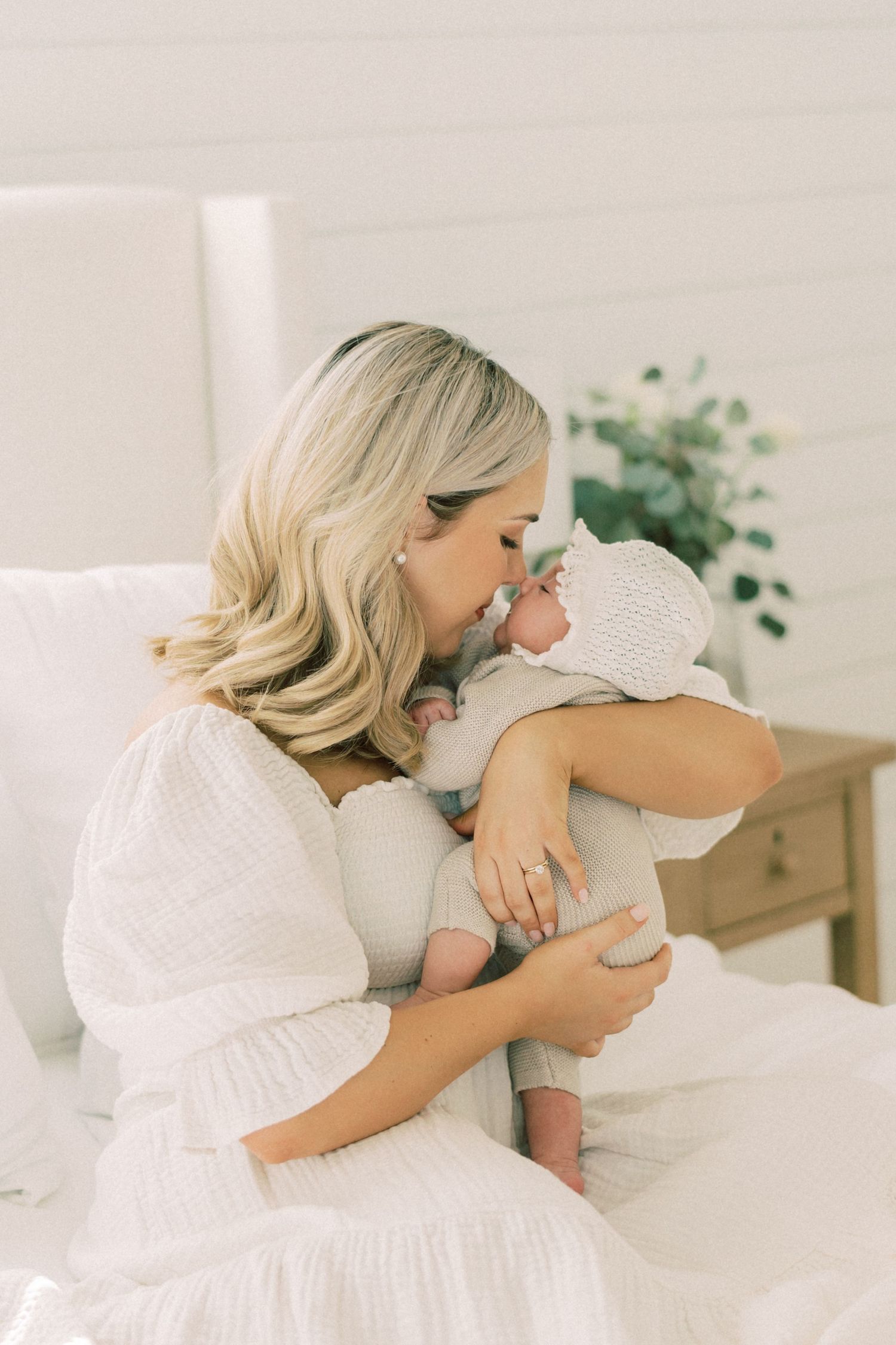 Tender moment shows a woman in white dress holding and kissing a small baby in a bright, airy room with houseplant.