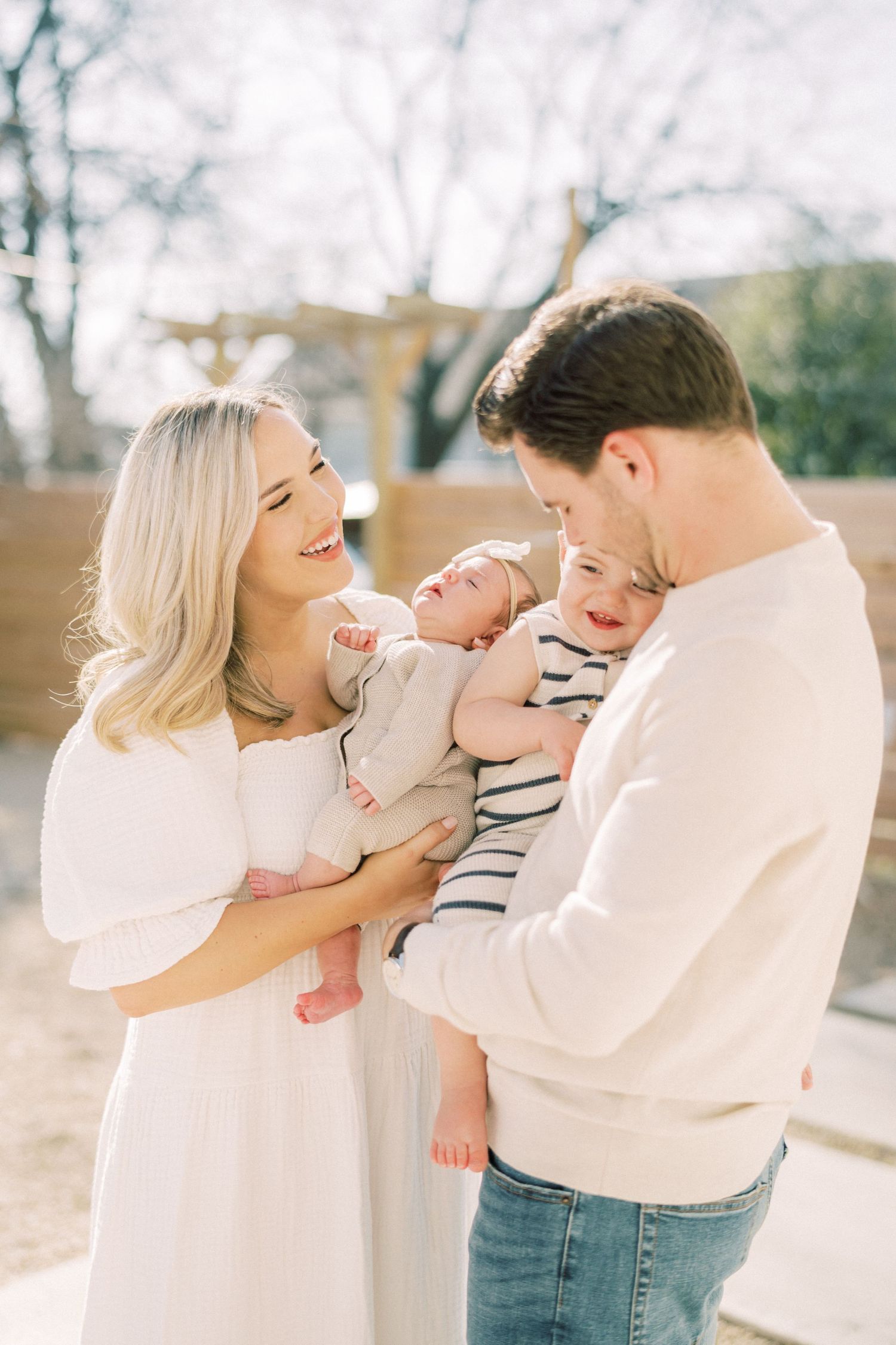 A tender family moment outdoors as parents in matching white sweaters share loving interactions with their young children.