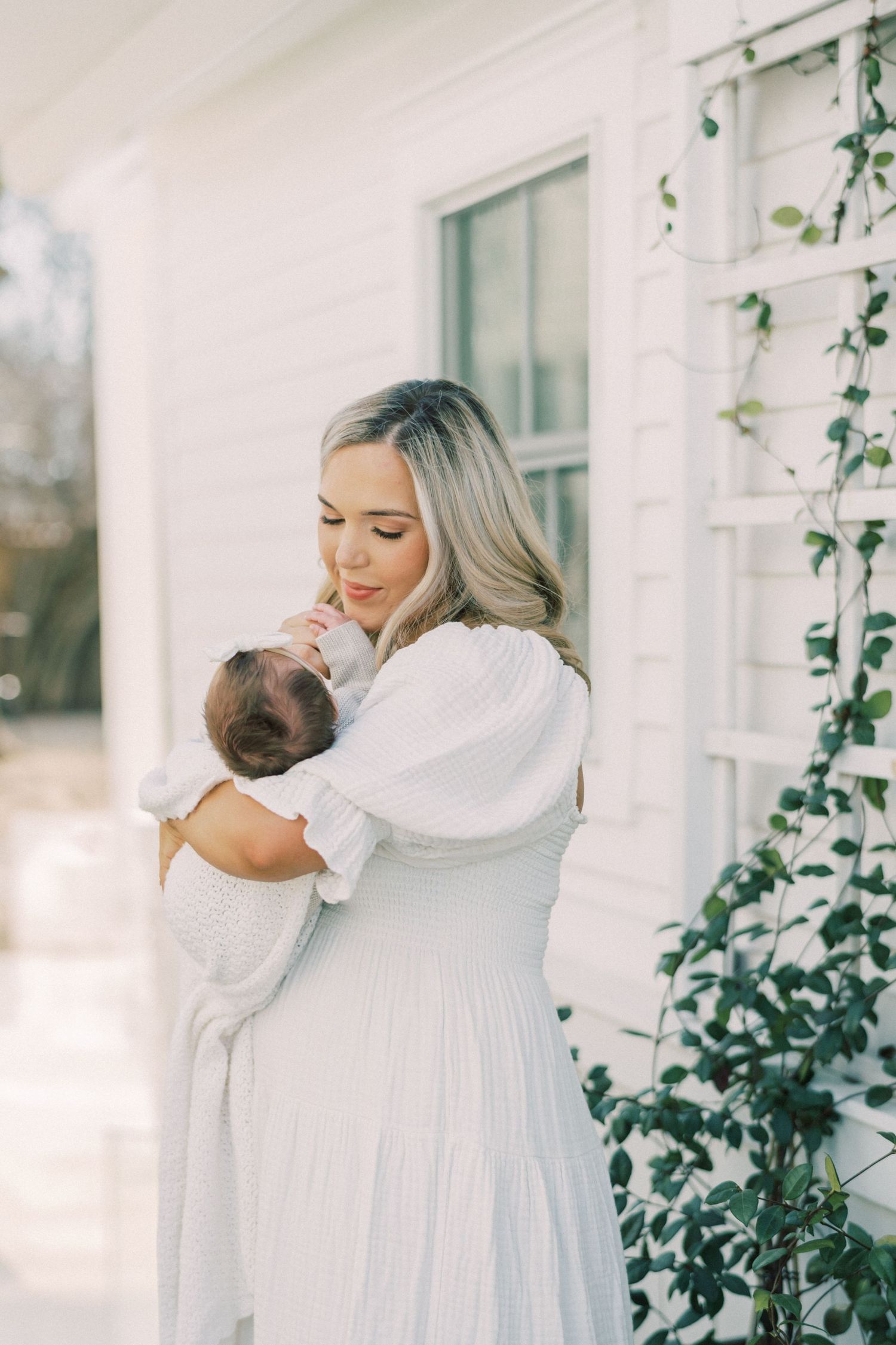 A woman in a flowing white dress holds a baby beside a white house with climbing vines.