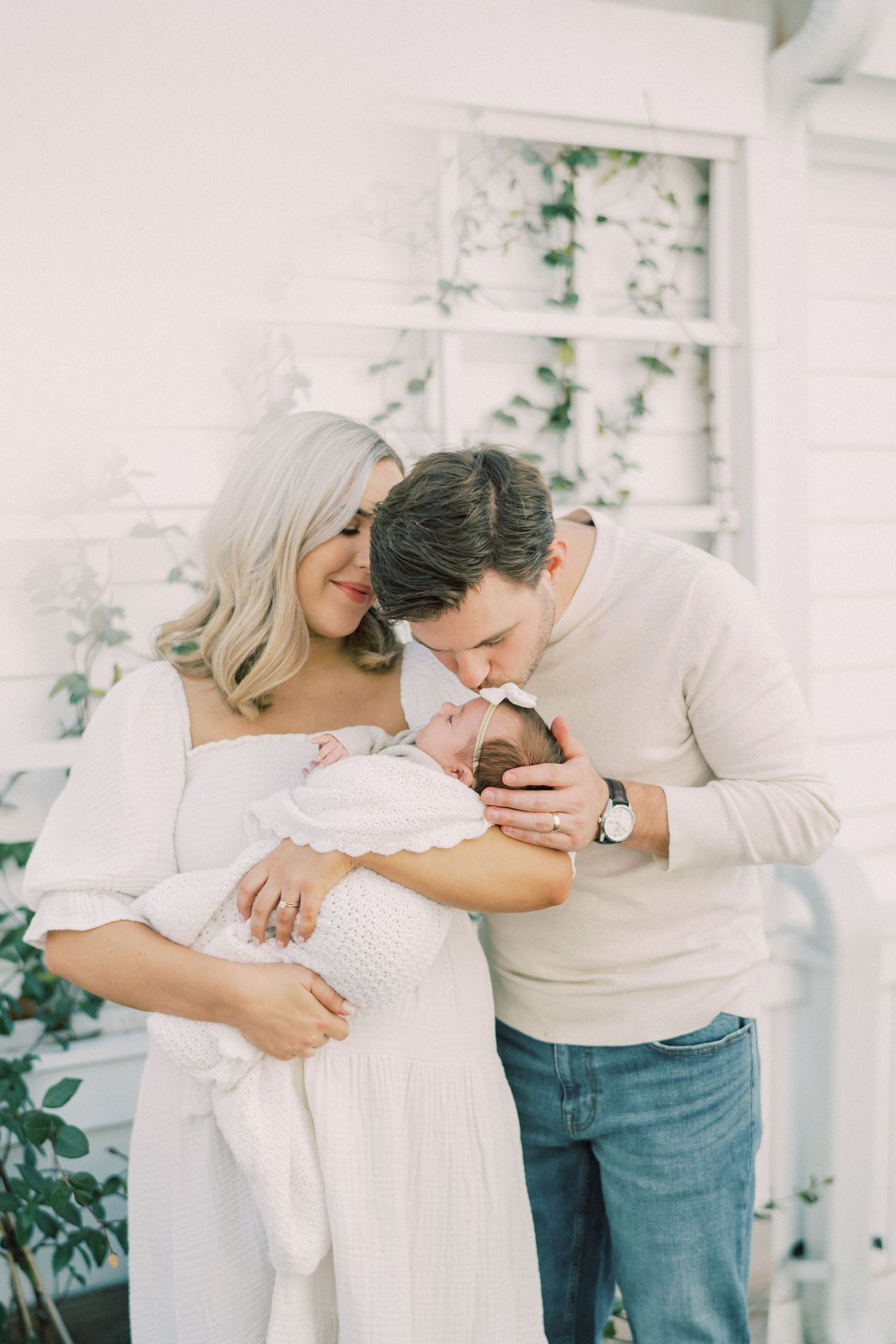 Modern family in matching white outfits sharing tender moment with newborn baby in outdoor garden setting.