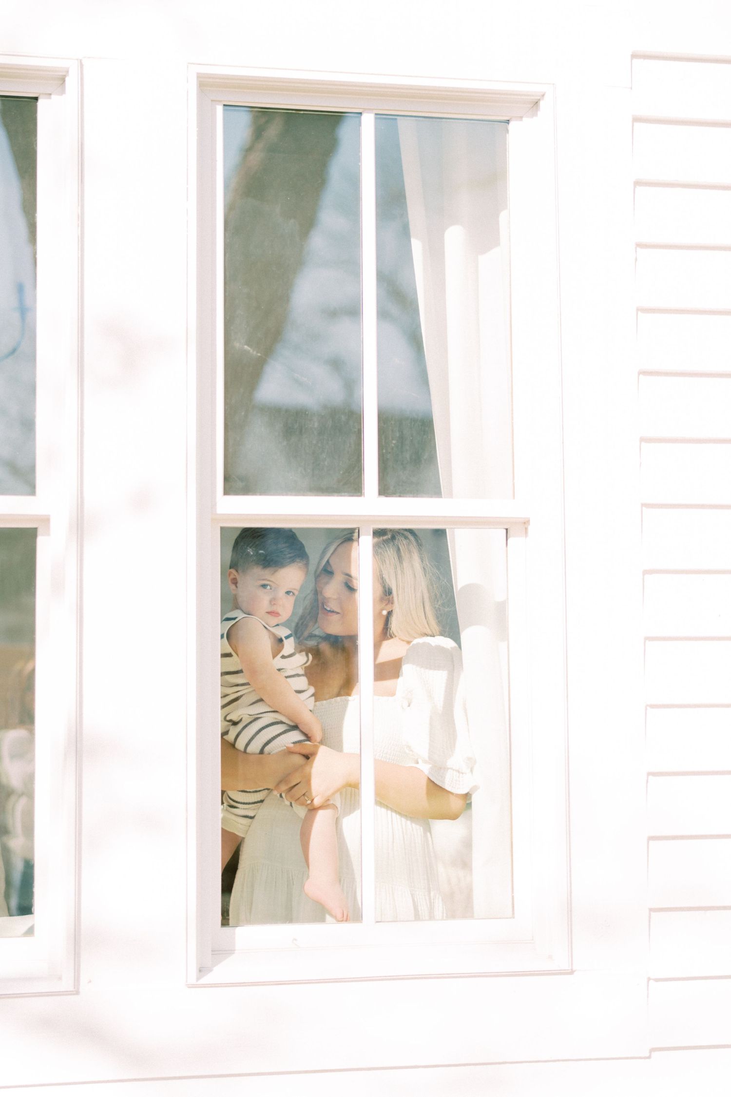 Through a bright window pane, two figures tenderly share a moment while holding a small baby inside a white-walled room.