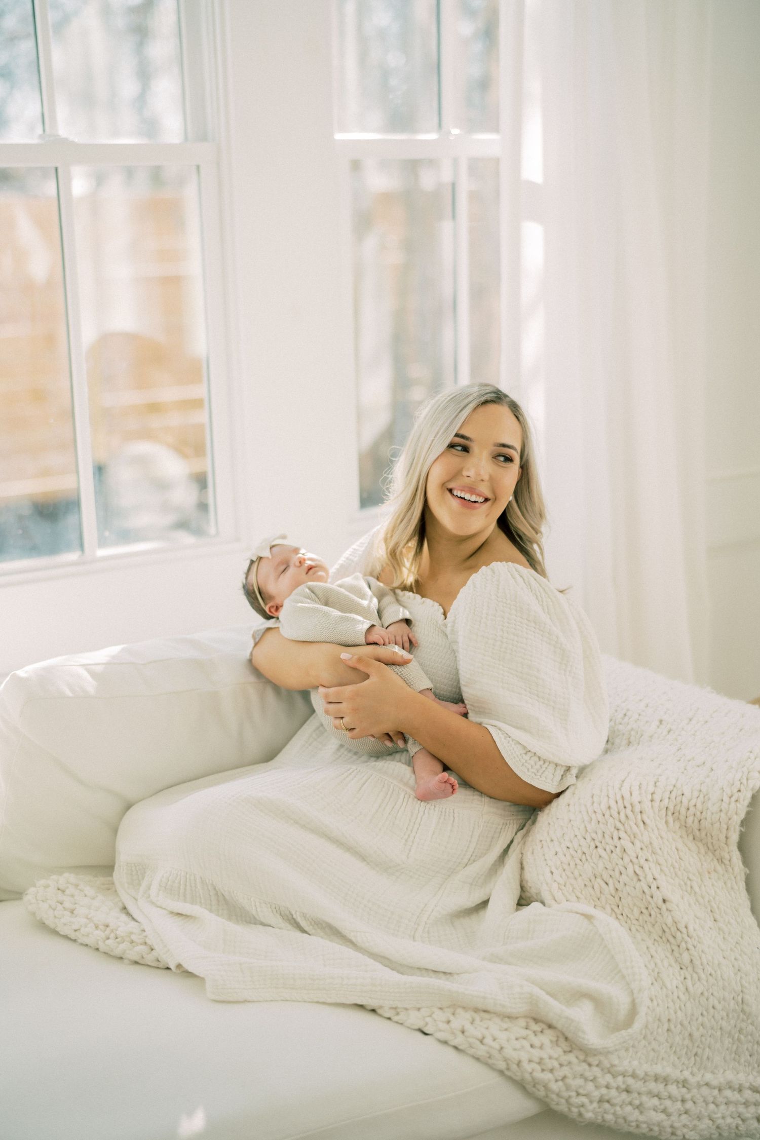 A parent in a white dress sits on a couch holding a sleeping newborn baby, with natural light streaming through windows.