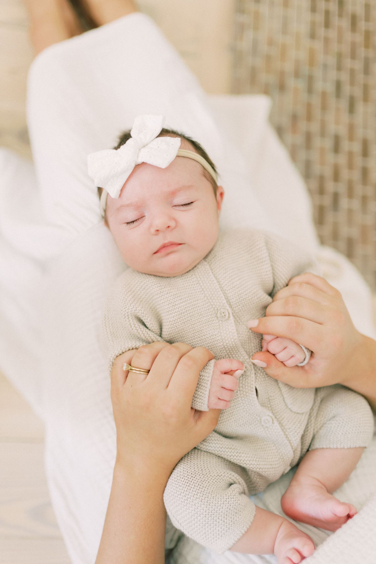Sleeping baby wearing a white bow headband and knit romper lying on soft white fabric while held by caring hands.