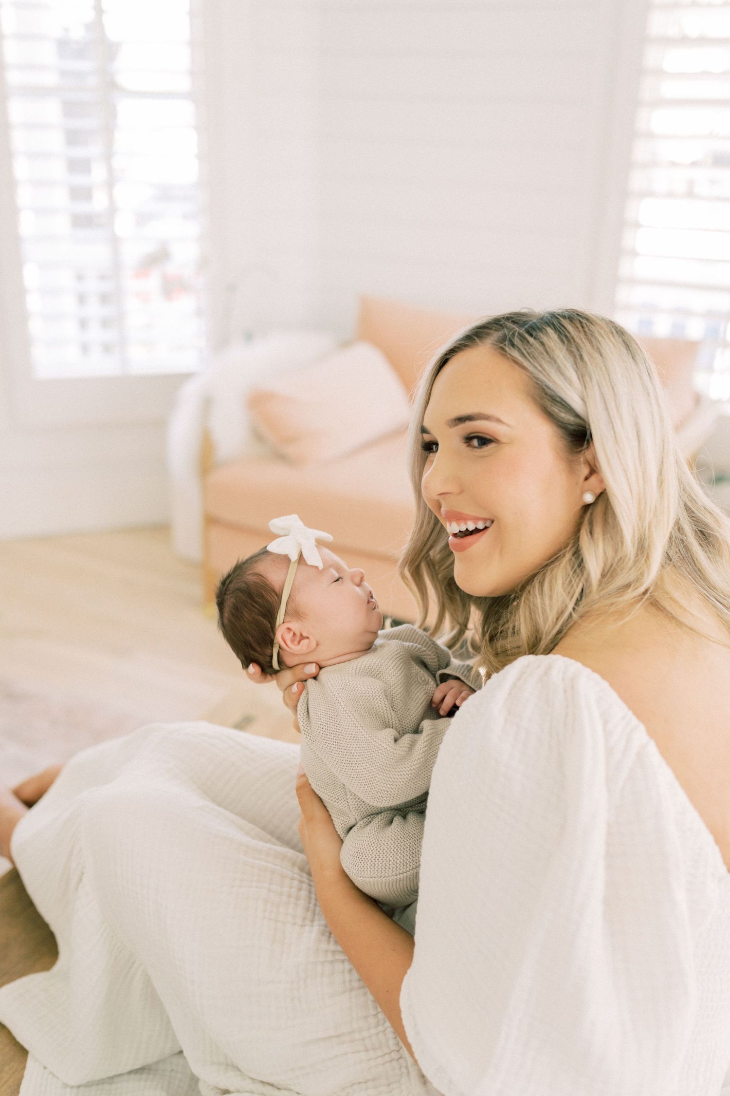 A woman in a white dress sits with a newborn baby wearing a grey onesie in a bright, minimalist setting.