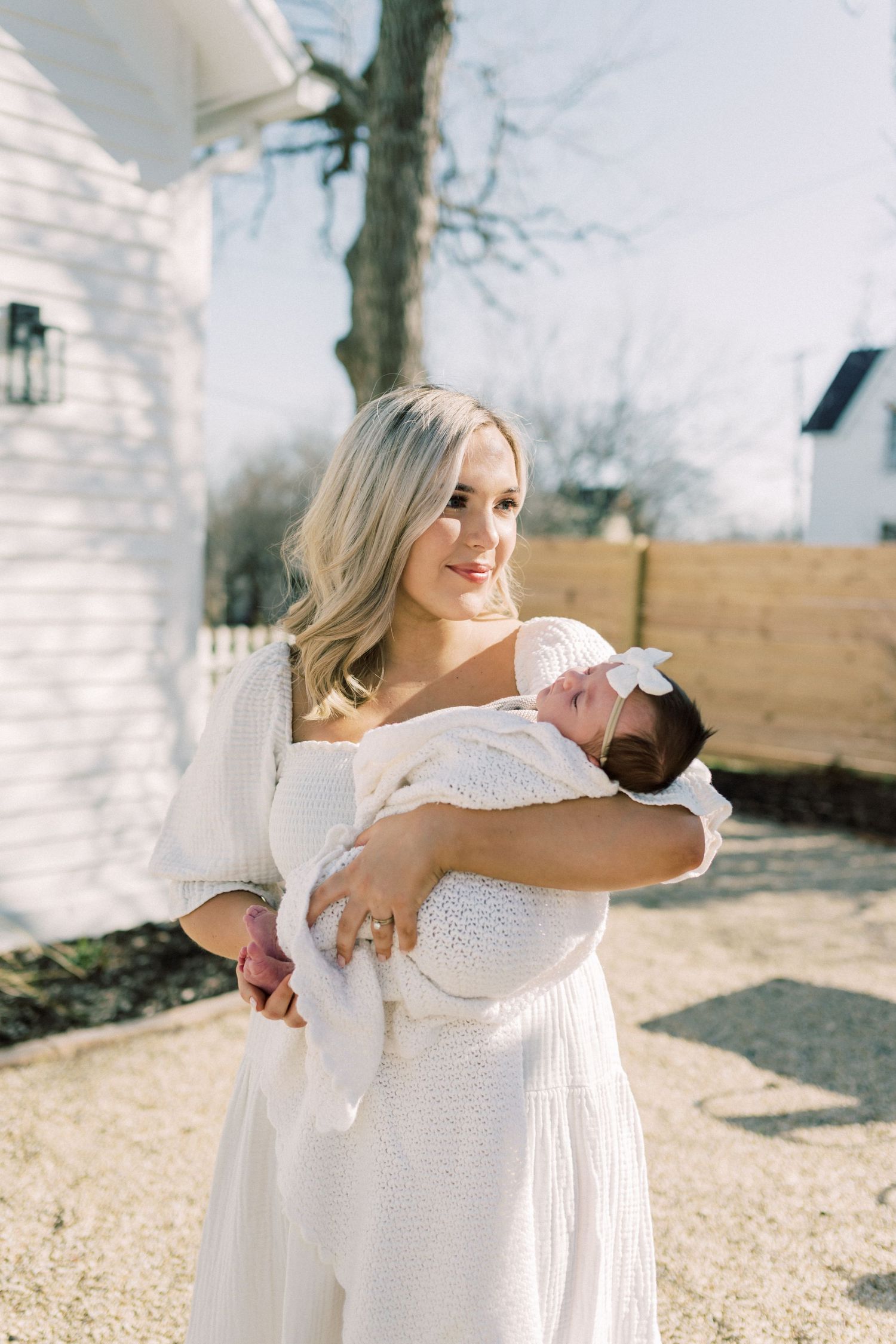 A woman in a flowing white dress holds a small baby wrapped in blankets while standing outside a house on a sunny day.