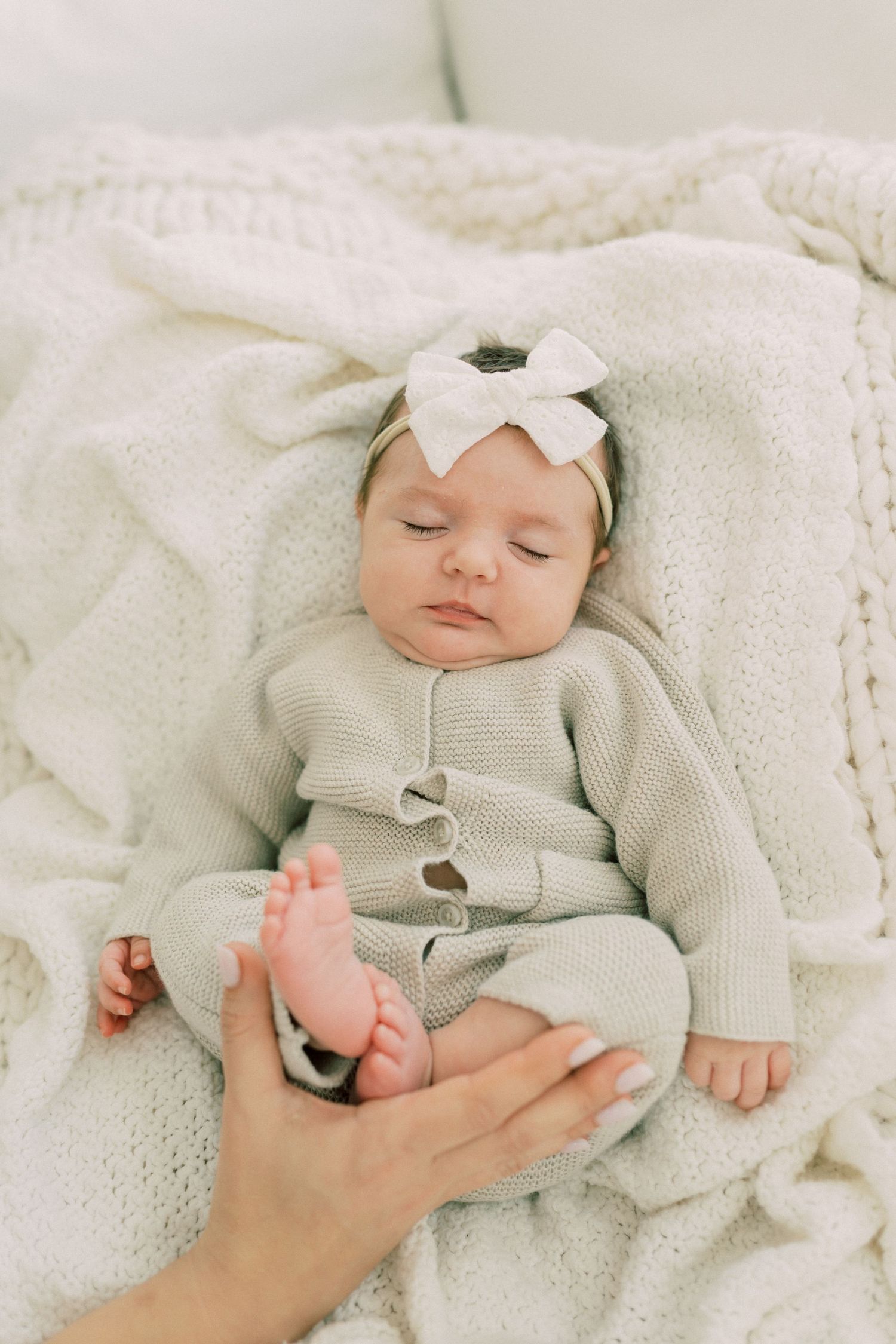 A sleeping baby wearing a white bow headband and gray cardigan lies on soft white blankets while a hand touches their foot.