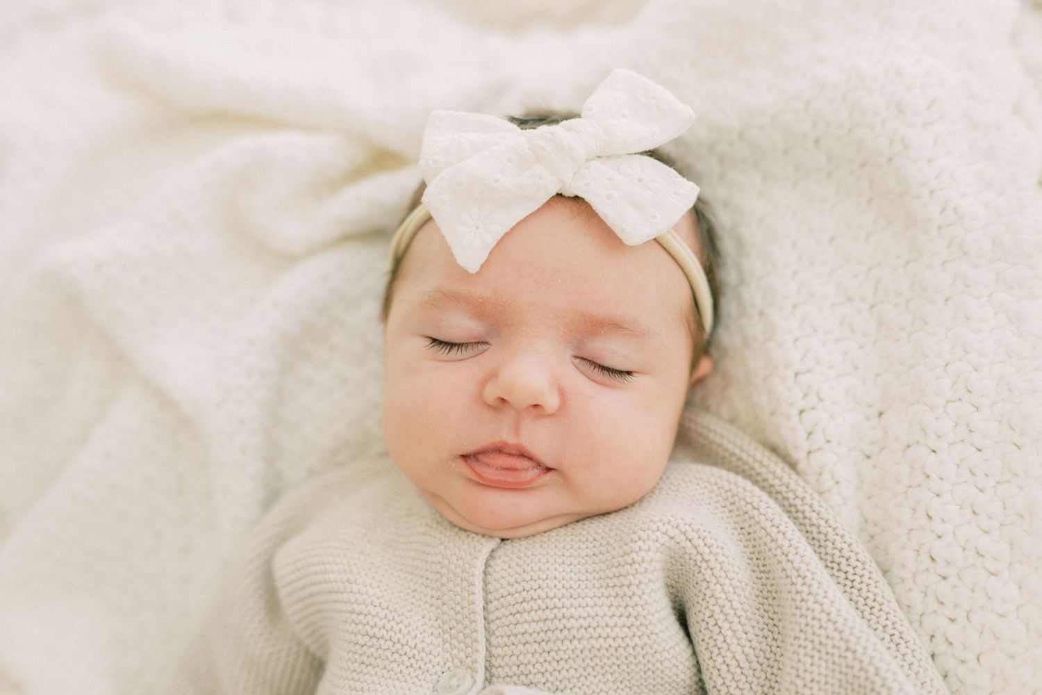 Sleeping baby wearing white knit sweater and bow headband rests on soft white blanket.