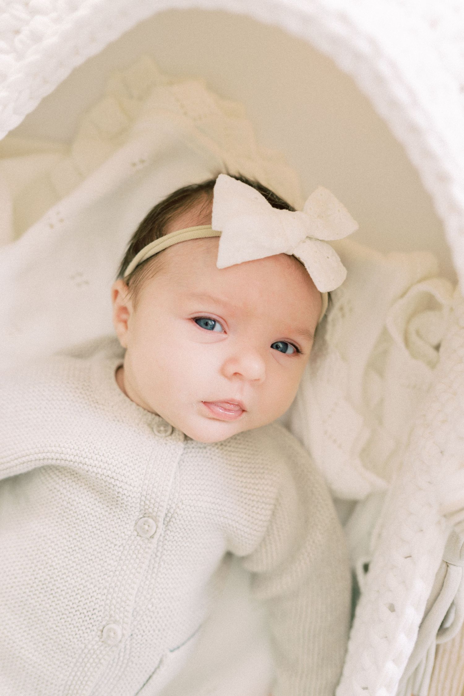 Baby wearing a white knit sweater and white bow headband rests on cream colored blanket.