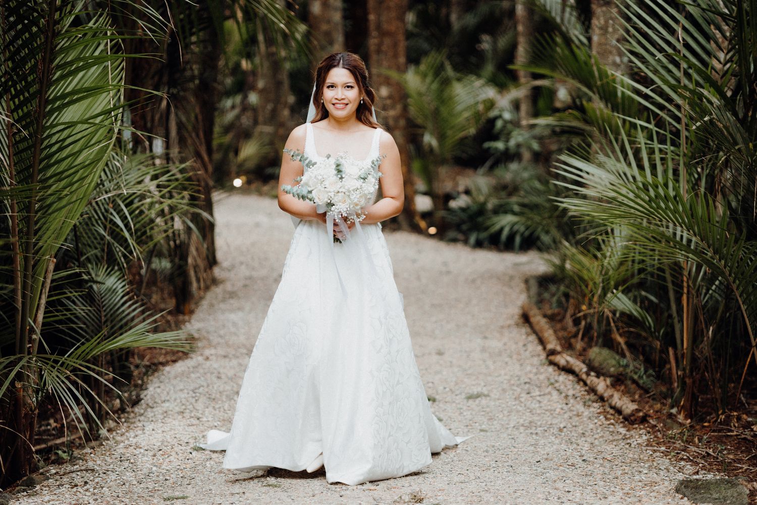 Beautiful bride in white off-shoulder wedding gown poses on garden path captured by Apertured Media Auckland Wedding Photographer.
