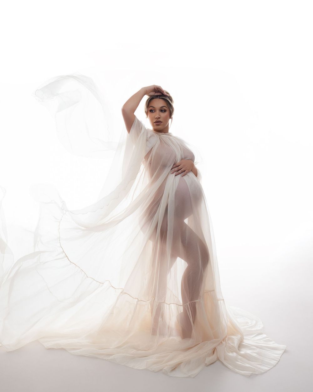 An ethereal maternity photo featuring a flowy sheer white gown against a light background.