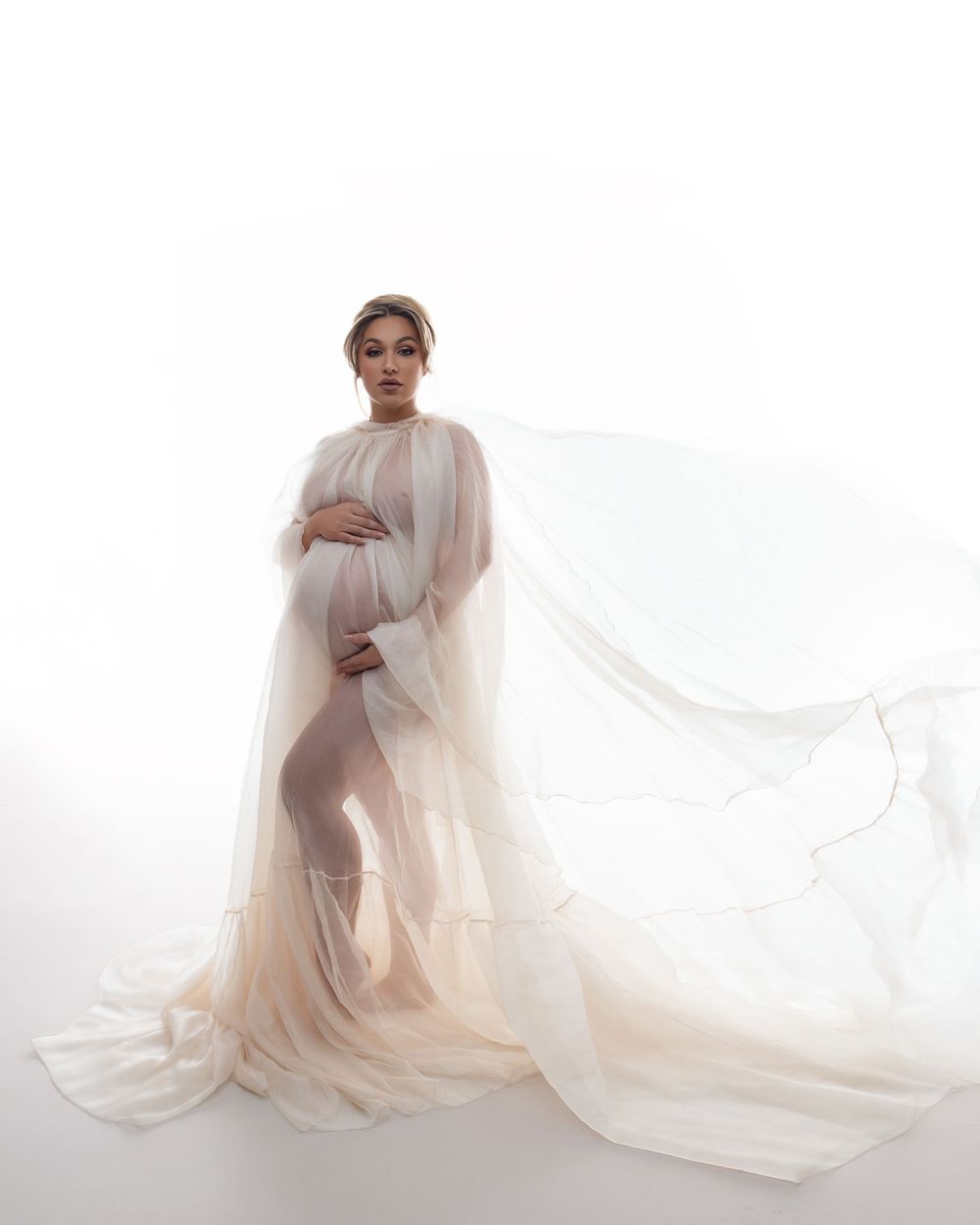 Ethereal maternity portrait with flowing sheer fabric in soft cream tones against white background.