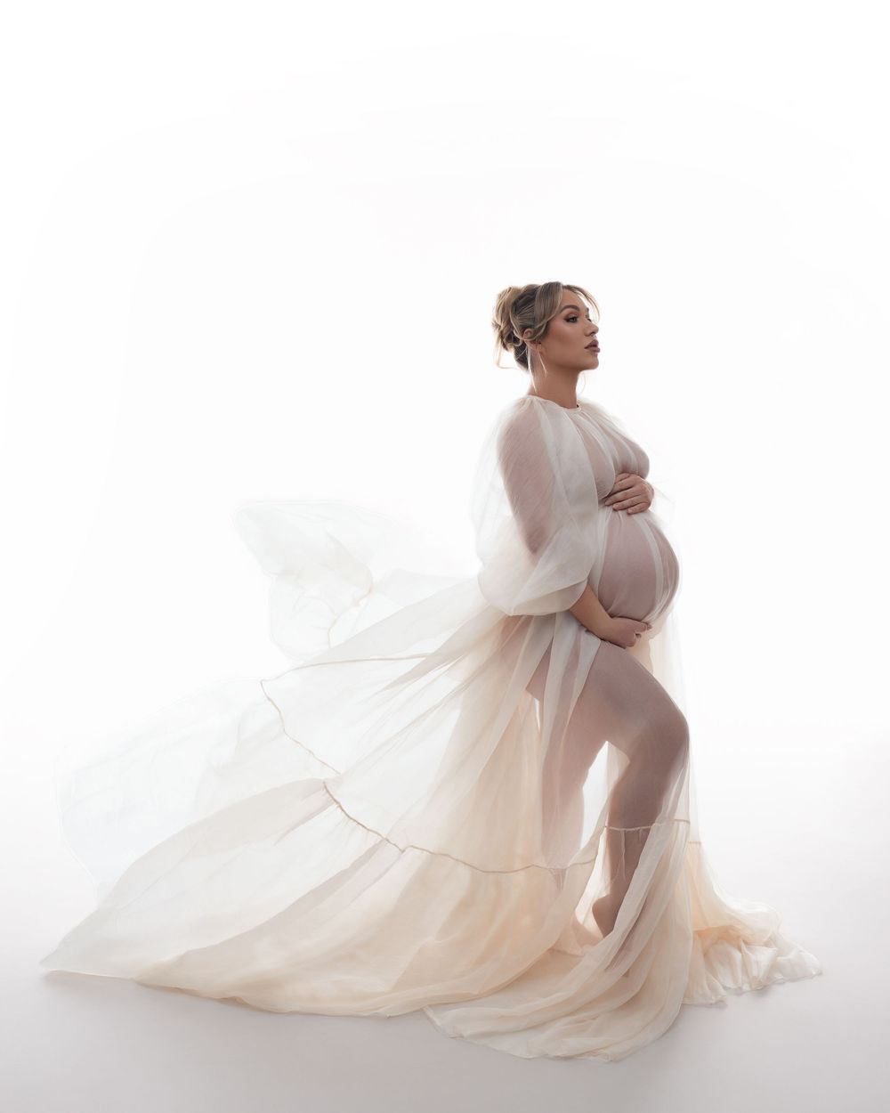 A flowing sheer cream dress elegantly drapes around an expectant mother in an ethereal maternity portrait.