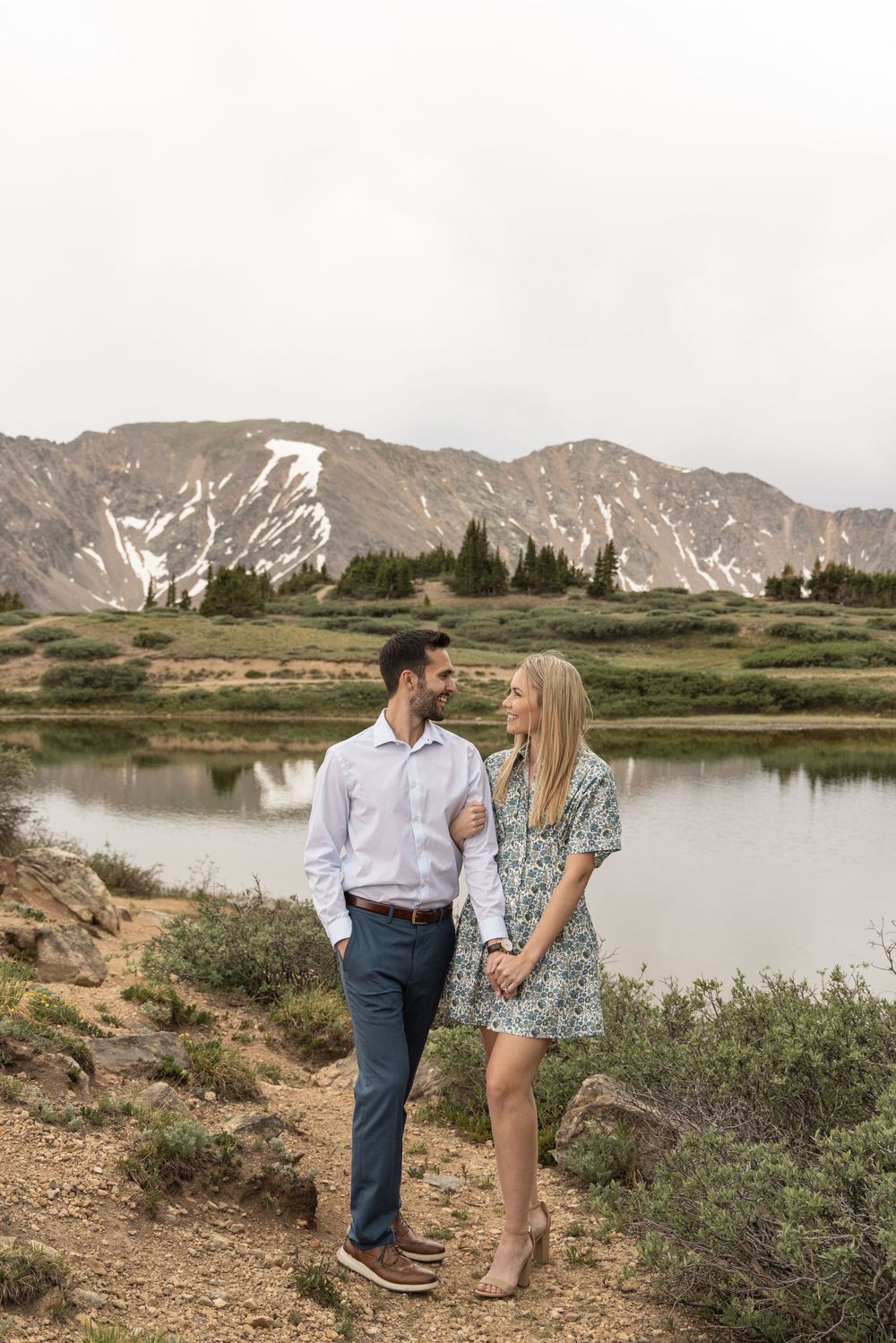 A couple shares a loving moment by a serene mountain lake with snow-capped peaks in the background.