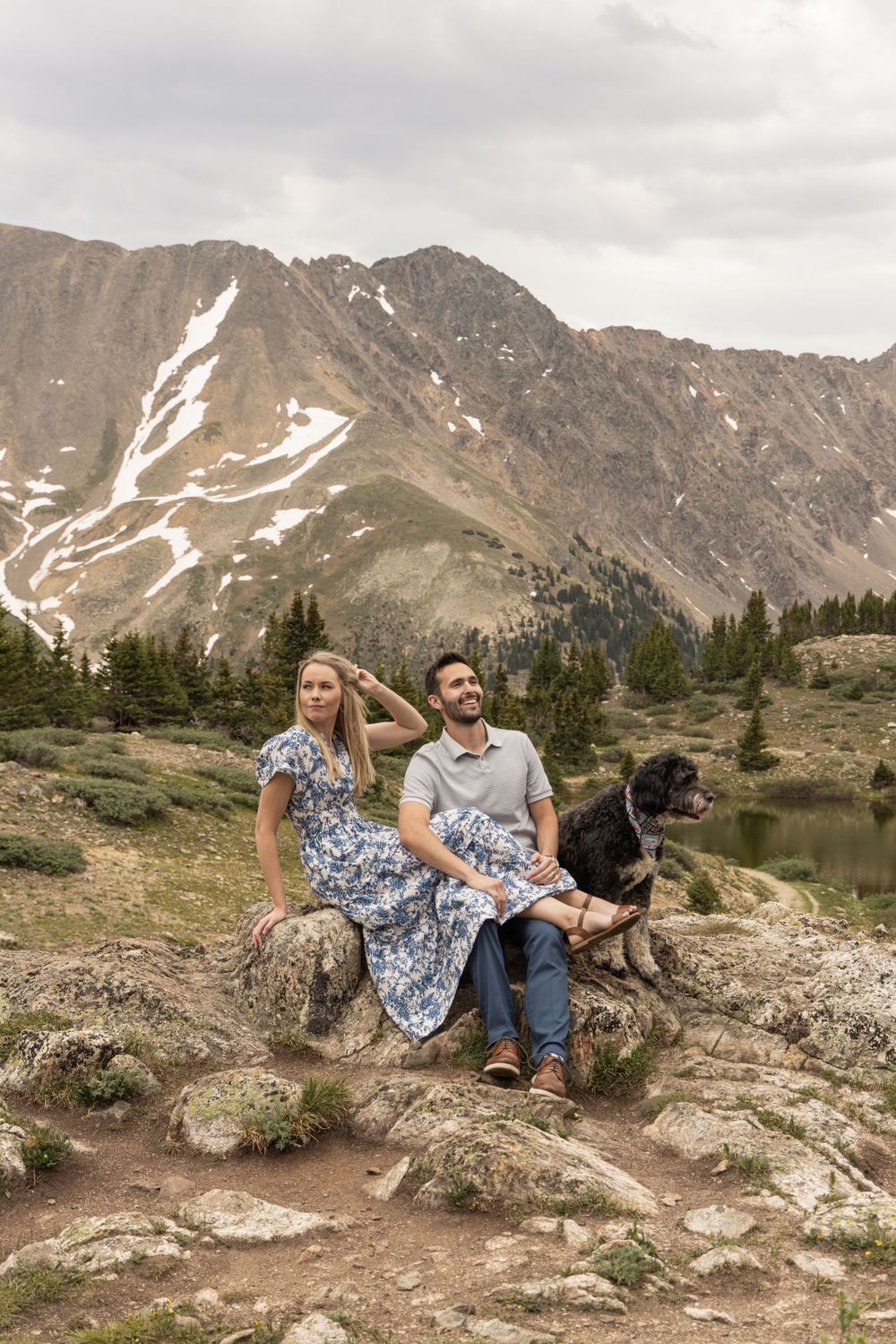 Couple and their dog sit on rocky outcrop with snow-capped mountains and alpine lake in Colorado wilderness backdrop at Loveland Pass.