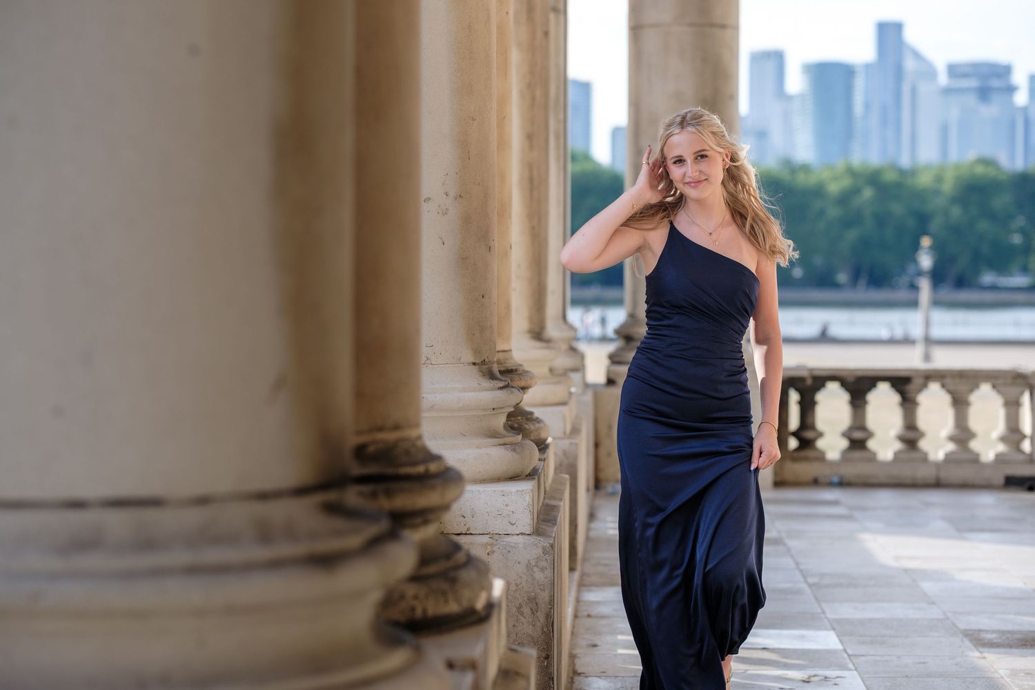 Woman in elegant navy blue evening gown poses by classical columns with city skyline in background.
