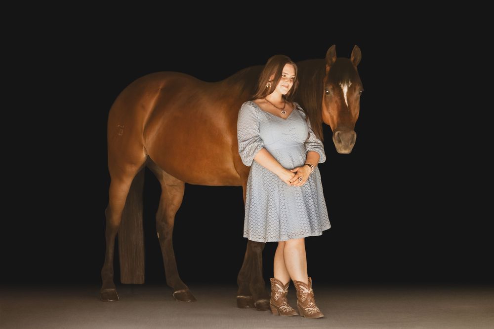 A young girl in a flowy gray dress stands beside a majestic bay horse against a dark background.