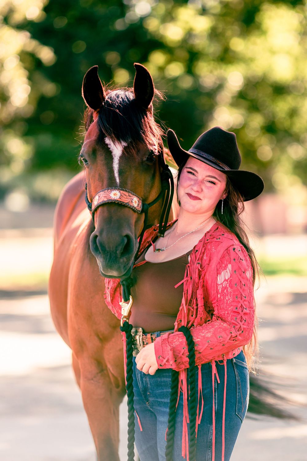 A bay horse stands with someone wearing a black cowboy hat and red lace top in an outdoor setting.