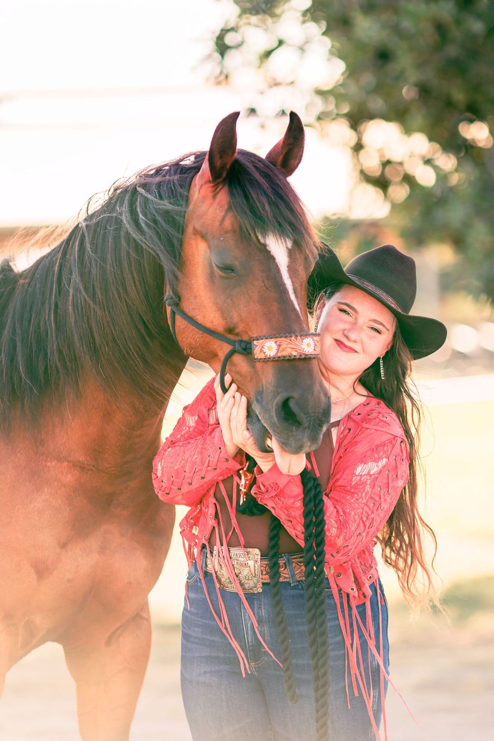 A heartwarming moment between a brown horse and someone wearing a coral top and hat in warm golden sunlight.
