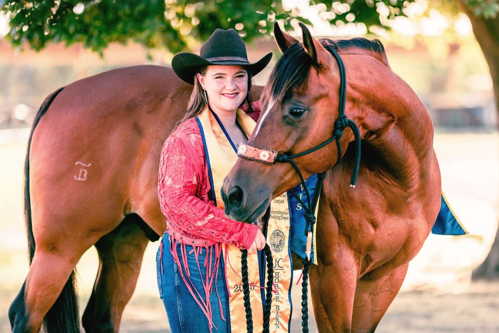 A rider in western attire stands beside a bay horse in an outdoor setting during golden hour.