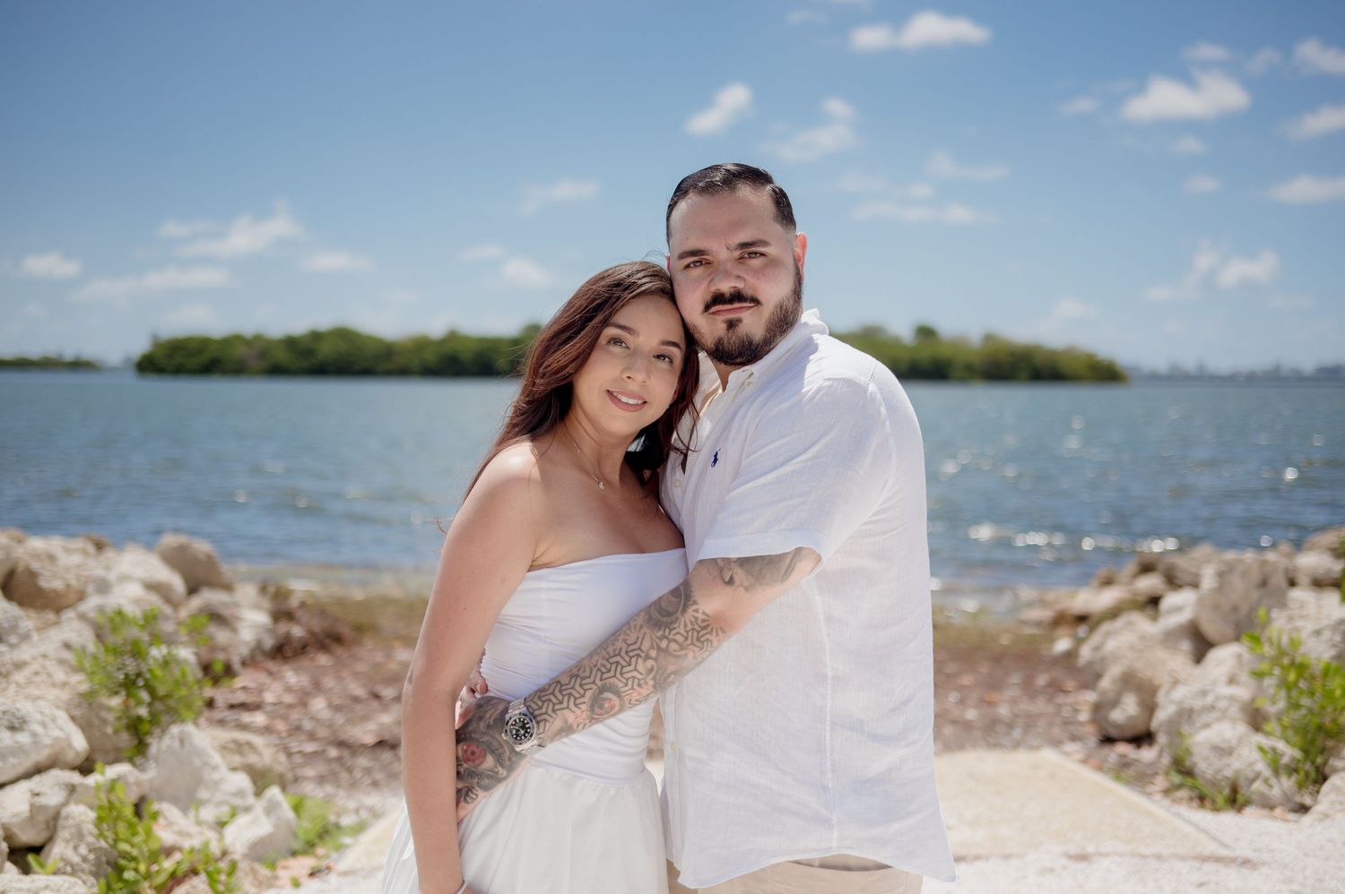 A couple poses together on a rocky shoreline with blue water and tropical islands in the background.