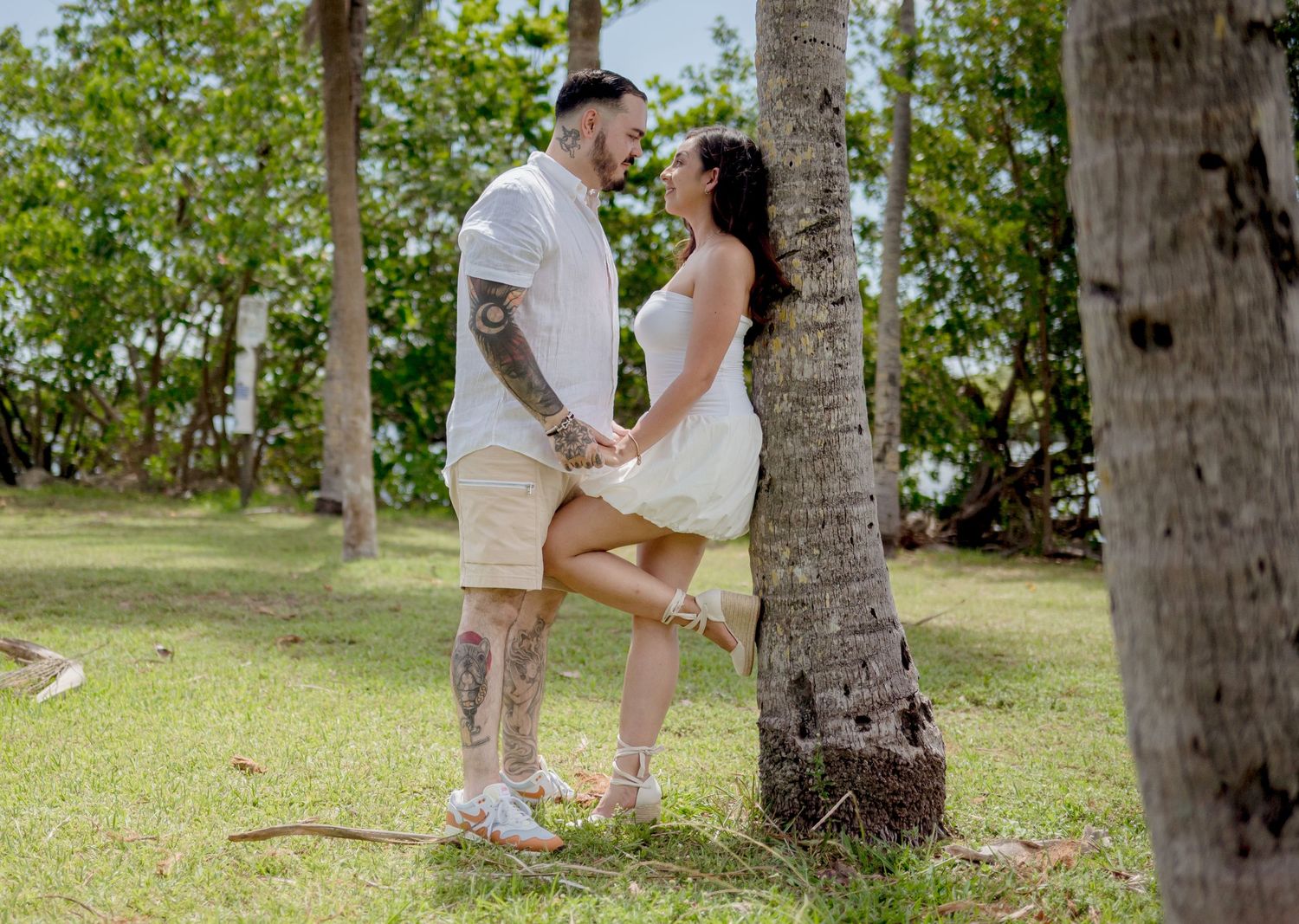 A couple in casual white attire shares a romantic moment beneath palm trees on a grassy lawn in a tropical setting.