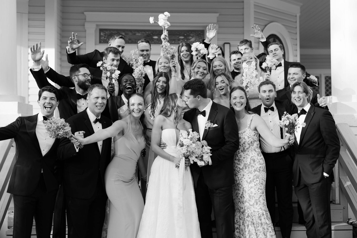 A black and white photo of a wedding party celebrating together on steps with raised glasses and bouquets.
