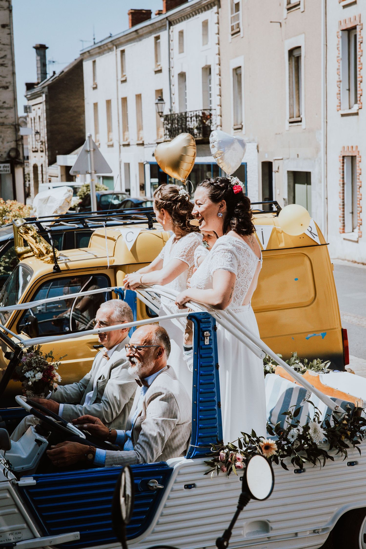 A vintage yellow car carries passengers through a quaint European street during a celebratory event.