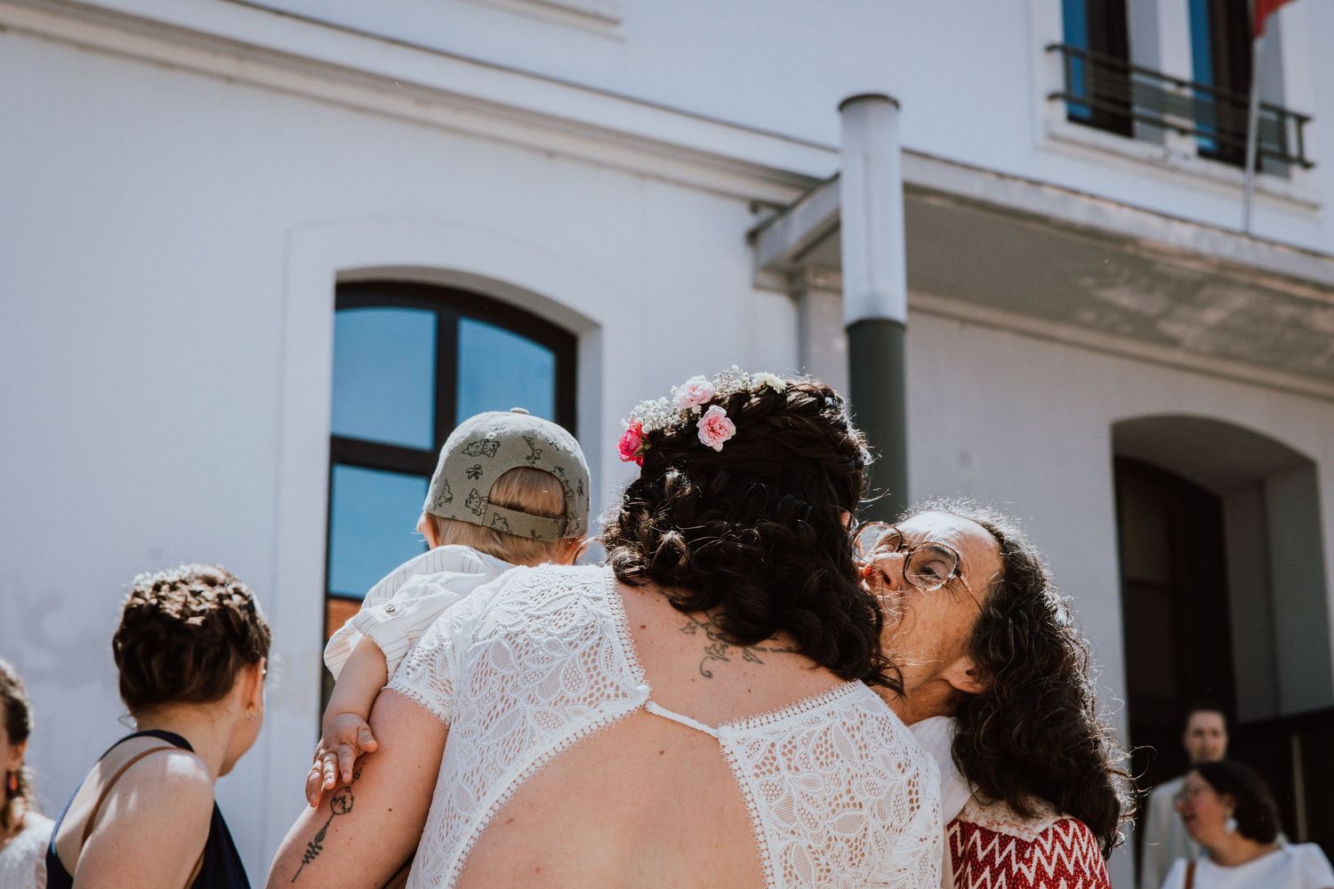 A joyful outdoor celebration features people in white lace dresses and floral crowns gathered at a building entrance.