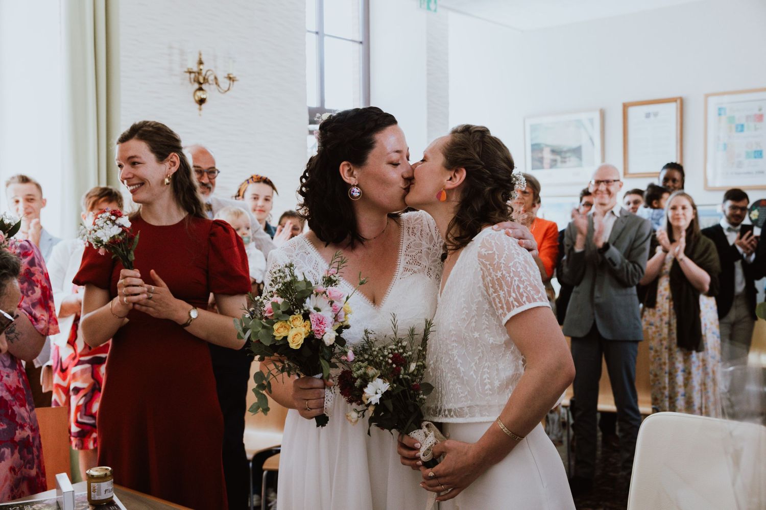 Two people in wedding dresses share a romantic kiss while holding bouquets during an indoor celebration.