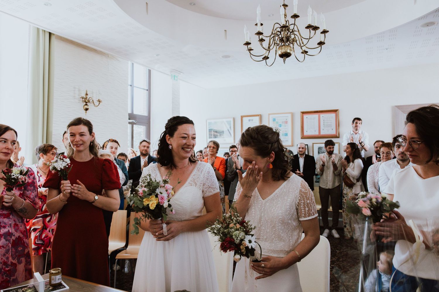 A joyous celebration inside a bright white room with a chandelier as guests gather for a special ceremony.