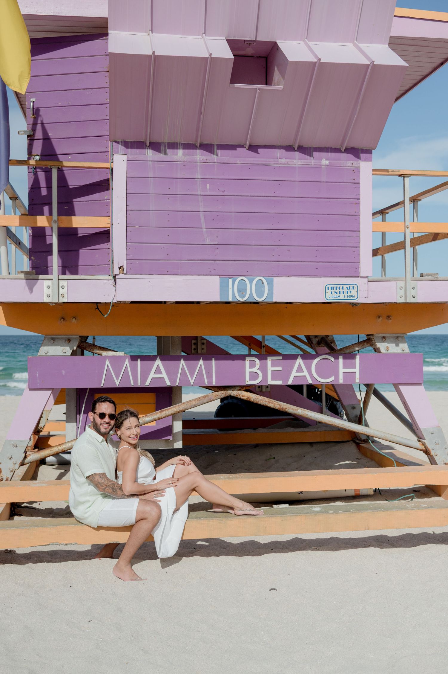 Person sits under purple Miami Beach lifeguard tower on sunny sandy beach during summer vacation.