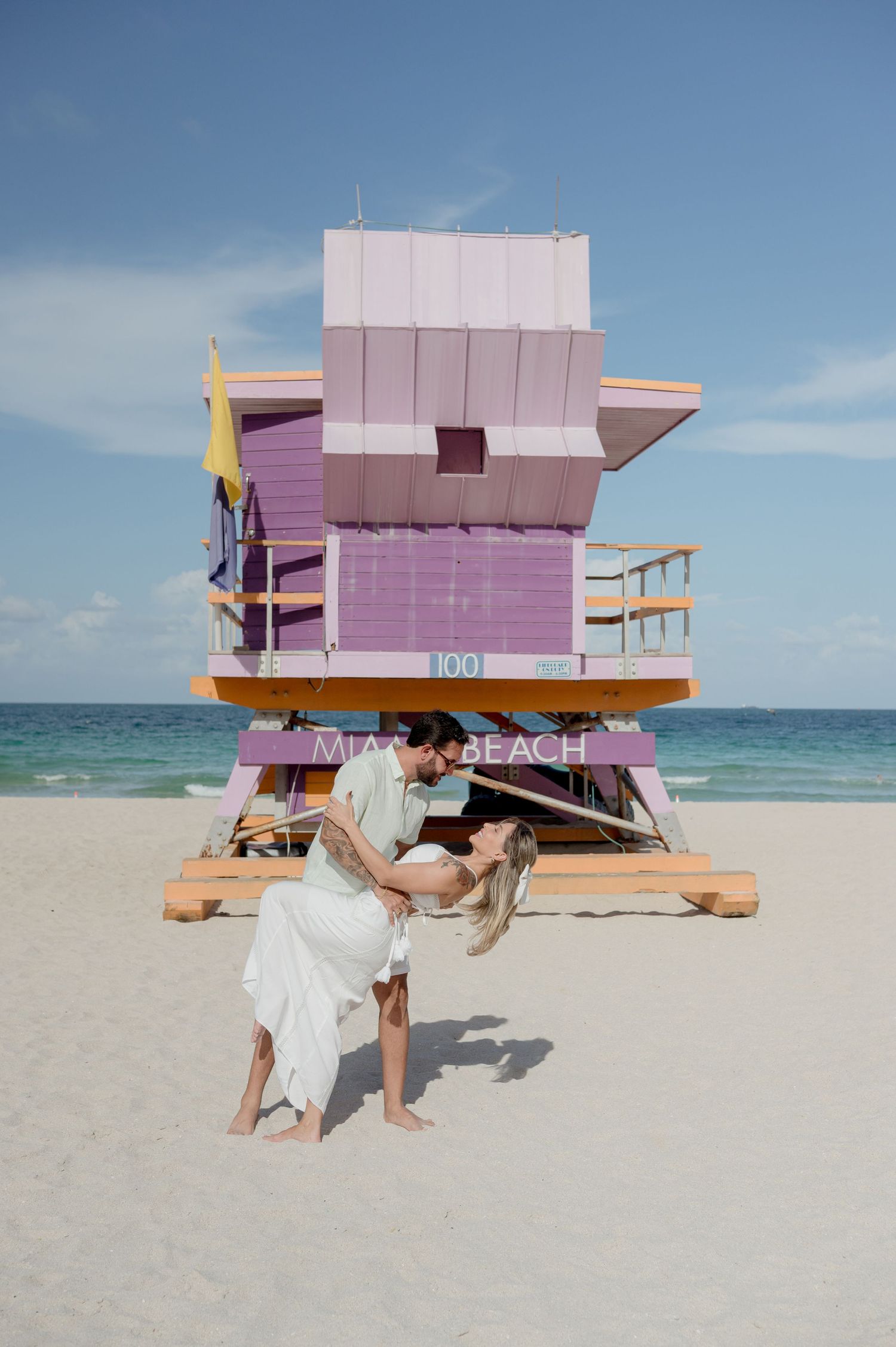 Two people posing in white outfits in front of a pink lifeguard tower on a sunny Miami Beach.