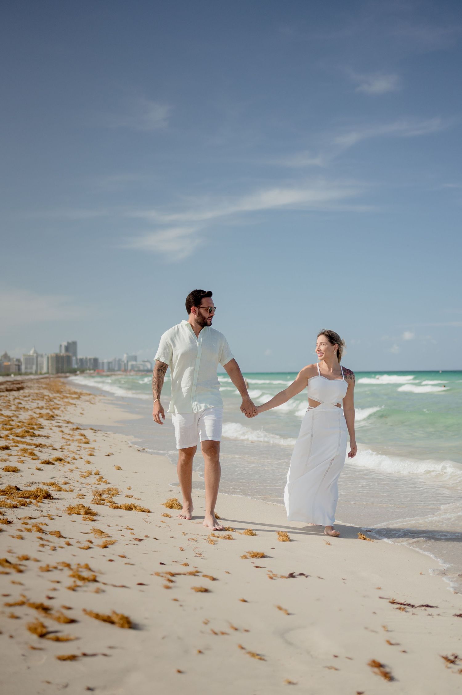 Two people walking along a scenic Miami Beach shoreline with waves and seaweed on a sunny day.