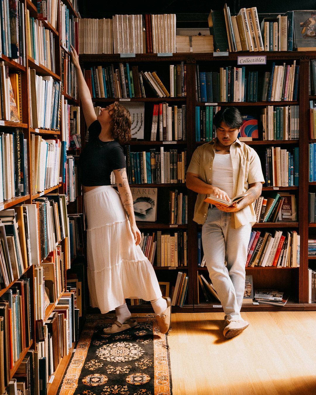 A woman reaching for books on high shelves in a cozy library with wooden bookcases with a man next to her.