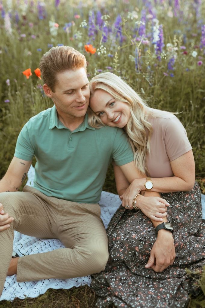 Romantic couple sits together among purple wildflowers and orange poppies in a scenic outdoor setting.