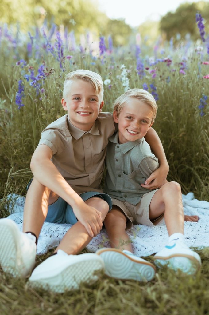 Two children sit together on a white blanket surrounded by purple wildflowers on a sunny summer day.
