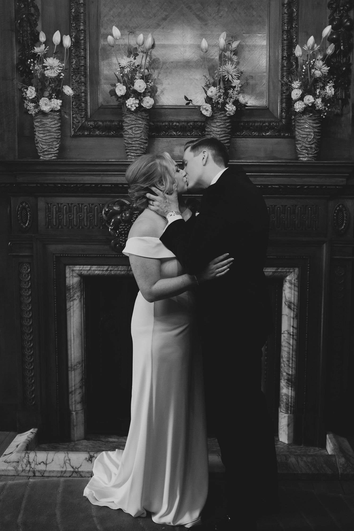 A romantic black and white wedding photo of a couple kissing in front of a fireplace adorned with floral arrangements.