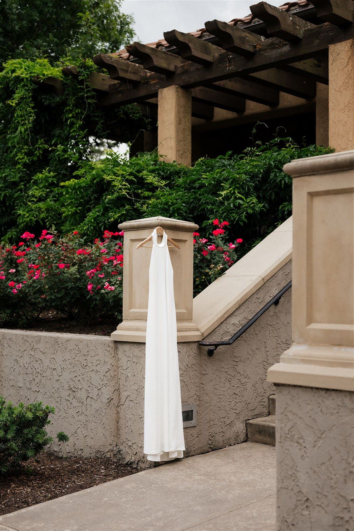 White wedding dress hanging on pergola wall with pink roses blooming in garden backdrop.