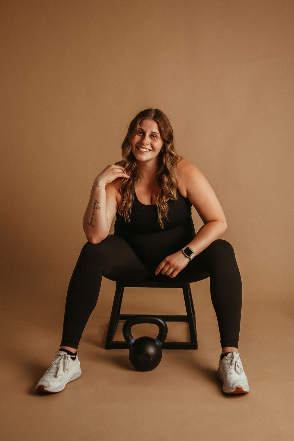Athletic figure in black workout attire sits on stool with kettlebell in studio against warm brown backdrop.
