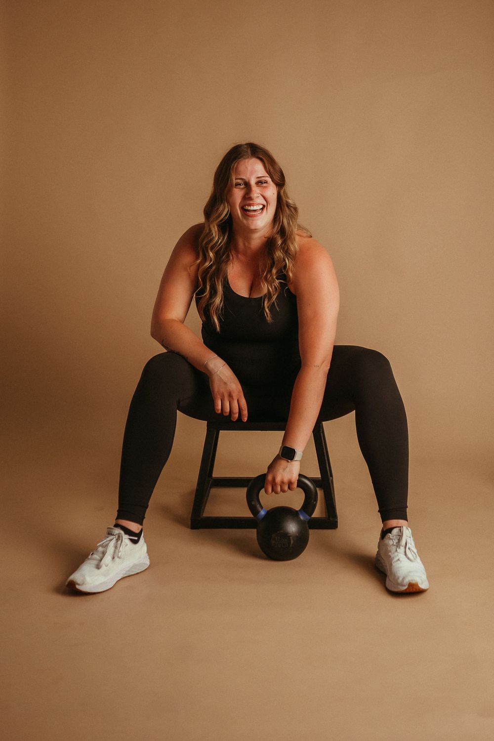 A seated athlete in black workout attire and white sneakers rests their hand on a kettlebell against a brown backdrop.