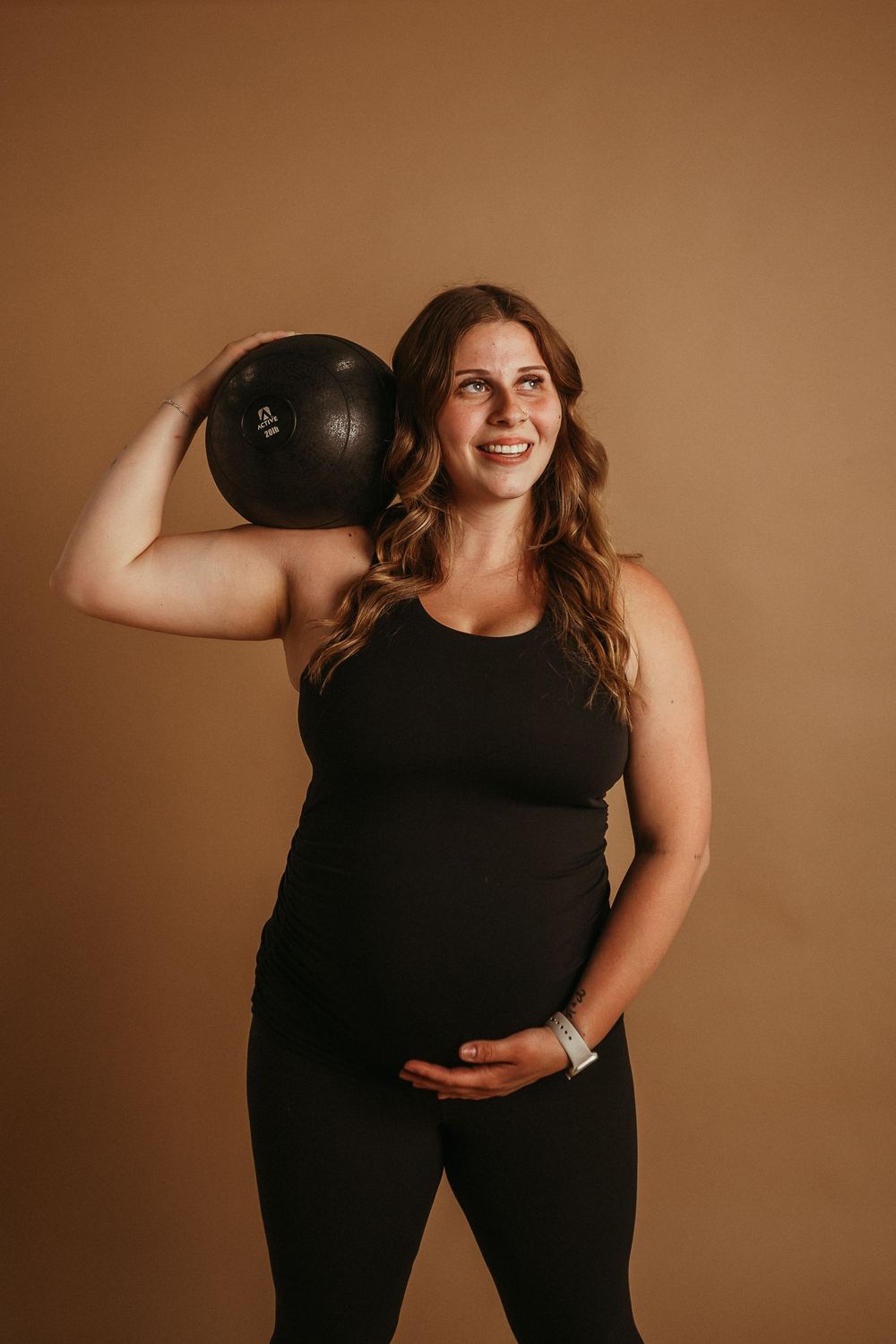 A pregnant person in black athletic wear holds a heavy workout ball while posing against a brown backdrop.